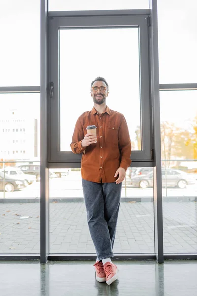 Overjoyed successful entrepreneur in eyeglasses, trendy shirt and jeans holding paper cup with coffee while standing at large windows in modern office, corporate lifestyle — Stock Photo