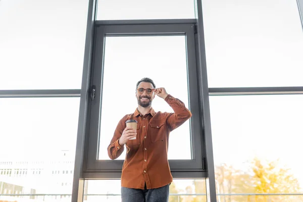 Low angle view of cheerful accomplished businessman in fashionable shirt standing with coffee to go near large office windows, adjusting eyeglasses and looking at camera — Stock Photo