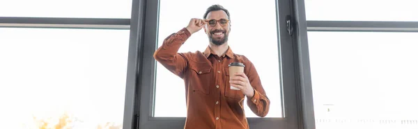 Low angle view of cheerful and accomplished businessman in stylish casual clothes holding coffee to go, adjusting eyeglasses and smiling at camera in office, banner — Stock Photo