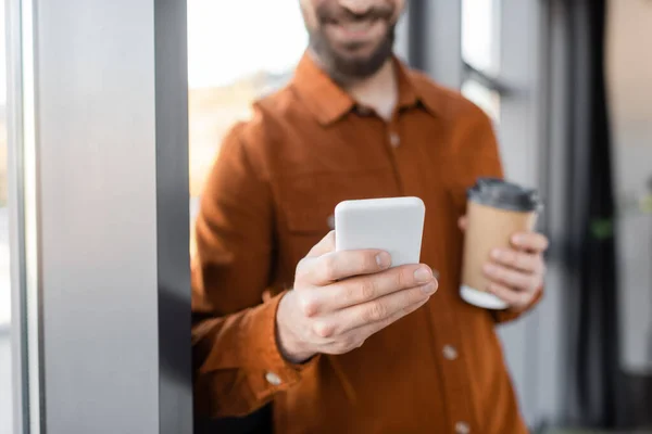 Частичный взгляд бородатого бизнесмена в стильной рубашке с сообщениями на мобильный телефон, стоя с кофе в одноразовой чашке в офисе, размытый фон — стоковое фото