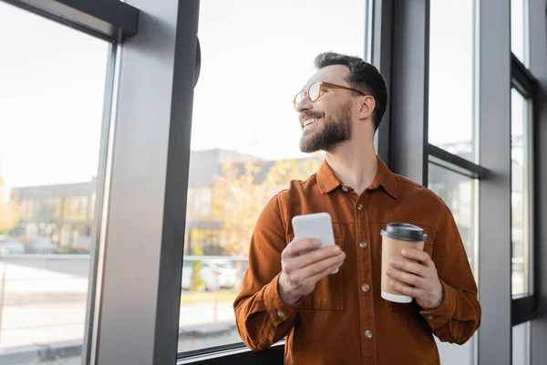 Веселый, успешный и бородатый бизнесмен в стильной рубашке и очках стоит со смартфоном и выпивкой на вынос возле окна в офисе и смотрит в сторону, корпоративный образ жизни — стоковое фото