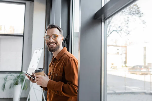 Good looking and joyful businessman in stylish shirt and eyeglasses standing with disposable cup and mobile phone near windows in office and looking at camera — Stock Photo