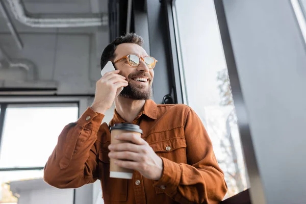 Hombre de negocios barbudo con gafas de moda y camisa de pie cerca de la ventana de la oficina con bebida para llevar en una taza de papel, sonriendo y mirando hacia otro lado sobre un fondo borroso - foto de stock