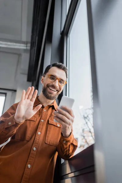 Low angle view of fashionable and positive manager in shirt and eyeglasses holding mobile phone and waving hand during video call near window in office, blurred foreground — Stock Photo
