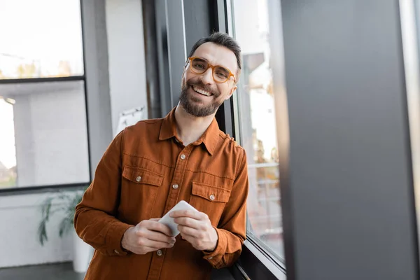 Empresário realizado e carismático em óculos elegantes e camisa de pé com telefone celular perto da janela e sorrindo para a câmera no escritório, foreground borrado — Fotografia de Stock