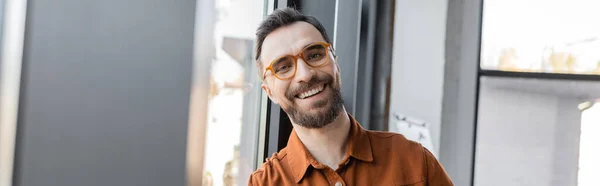 Portrait of charismatic and joyful businessman in trendy eyeglasses and shirt smiling at camera while standing near grey wall in modern office, banner — Stock Photo