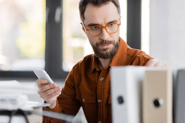 Homem de negócios concentrado e sério em óculos elegantes e camisa segurando telefone celular e olhando para pastas borradas enquanto trabalhava na produtividade do negócio no escritório — Fotografia de Stock