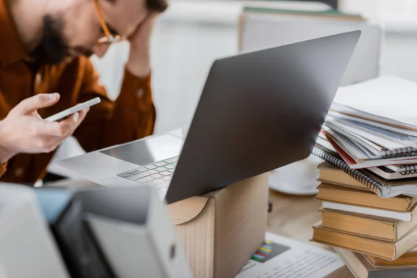 Exhausted manager with bowed head holding smartphone near laptop on carton box, folders and pile of books and notebooks at workplace in office, blurred background — Stock Photo