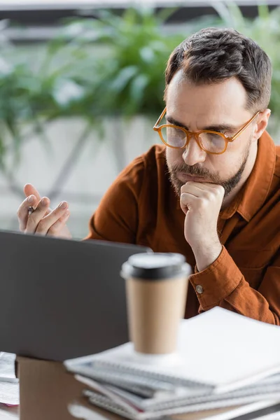 Homem de negócios cansado e pensativo em óculos elegantes sentados perto do laptop na caixa de papelão, pilha de cadernos e café para ir ao resolver o problema em primeiro plano desfocado no escritório — Fotografia de Stock
