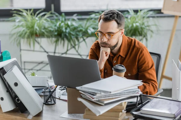 Deep in thought businessman puffing cheeks while solving problem near laptop on carton box, takeaway drink, pile of books and notebooks, pen holder with pencils and pens near scissors on desk — Stock Photo