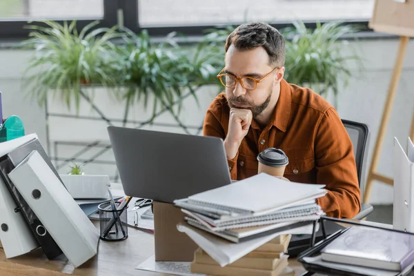 Profondément dans la pensée homme d'affaires gonflant les joues tout en résolvant le problème près d'un ordinateur portable sur la boîte en carton, boisson à emporter, pile de livres et de cahiers, porte-stylo avec crayons et stylos près des ciseaux sur le bureau — Photo de stock