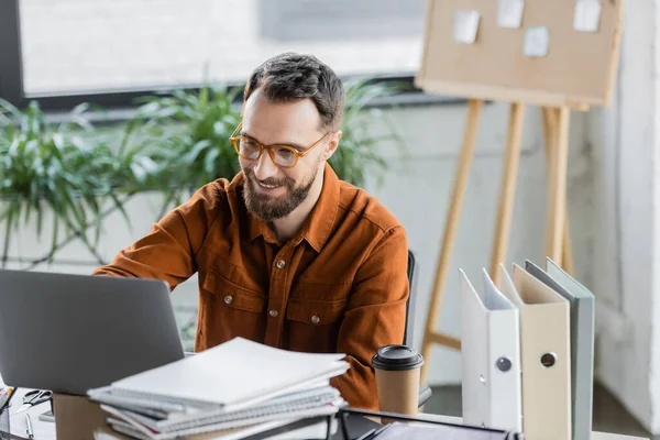 Successful executive manager in eyeglasses and shirt looking at laptop and smiling near pile of notebooks, folders, coffee to go and flip chart with sticky notes on blurred background — Stock Photo