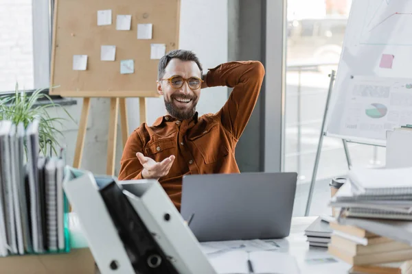 Cheerful bearded businessman in eyeglasses and shirt pointing with hand during video call on laptop at workplace with folders, books and notebooks while sitting next to corkboard with sticky notes — Stock Photo