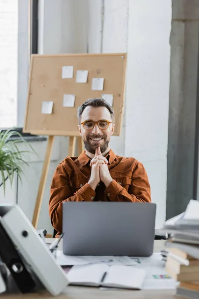Satisfied bearded businessman in trendy shirt and eyeglasses looking at laptop near blurred folders, notebooks, documents and flip chart with sticky notes on background in office — Stock Photo