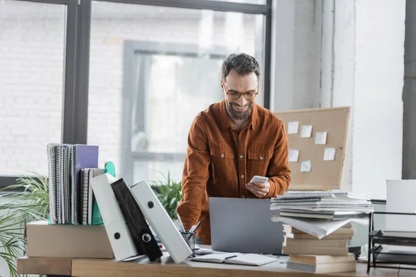 Uomo d'affari carismatico e barbuto in camicia elegante e occhiali guardando smartphone e sorridente vicino al computer portatile, un sacco di quaderni, cartelle e libri — Foto stock