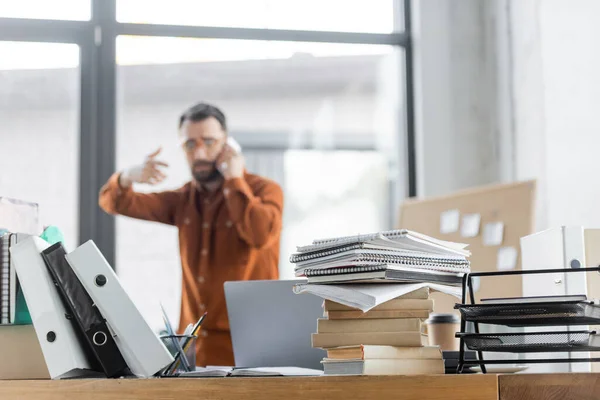 Selective focus of office desk with pile of books, notebooks, folders and laptop near busy businessman gesturing while talking on mobile phone and solving problem on blurred background — Stock Photo