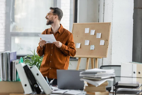 Positive successful entrepreneur holding document and looking away near cork board with sticky notes and work desk with laptop, folders, books and notebooks on blurred foreground — Stock Photo