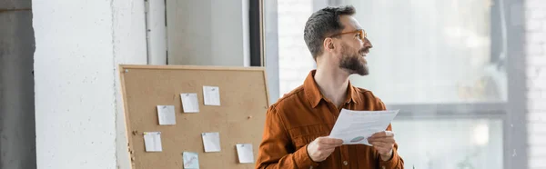 Accomplished and happy bearded businessman in eyeglasses and stylish shirt looking away near corkboard with attached sticky notes in modern office, banner — Stock Photo