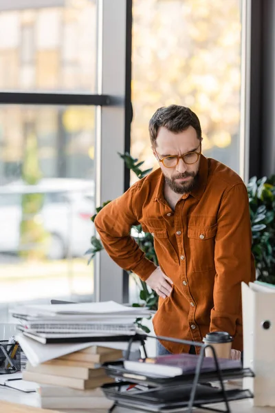 Pensive bearded businessman in eyeglasses standing with hand on hip and looking at plenty of books, notebooks and folders near paper cup on work desk in office — Stock Photo