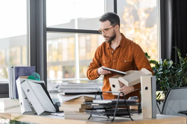 Serious bearded businessman in eyeglasses and shirt standing with carton box and notepad next to work desk with folders, pile of books, notebooks and coffee to go in office — Stock Photo