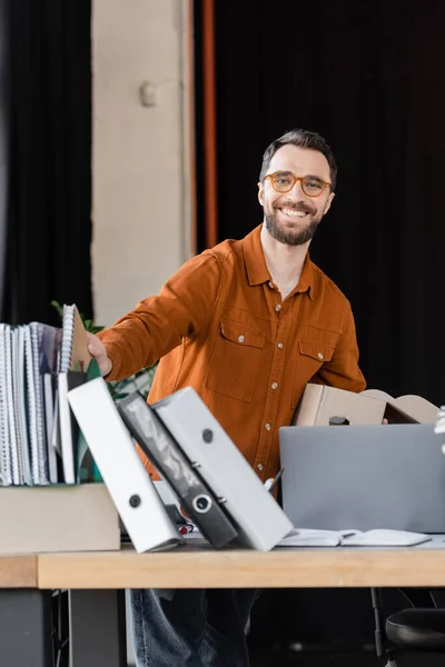 Cheerful manager in trendy shirt and eyeglasses holding carton box and smiling at camera next to workplace with notebooks, folders and laptop in modern office — Stock Photo