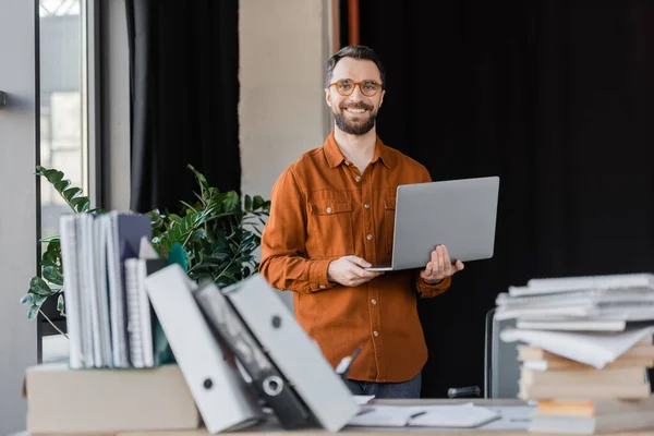 Happy successful businessman in stylish eyeglasses and shirt standing with laptop and looking at camera near pile of books, notebooks and folders on blurred foreground in office — Stock Photo
