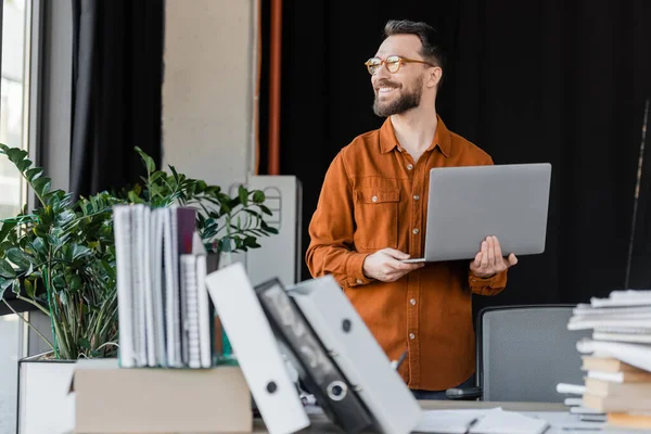 Positive and successful entrepreneur in eyeglasses and shirt standing with laptop and looking away near blurred pile of books, notebooks and folders on work desk in office — Stock Photo