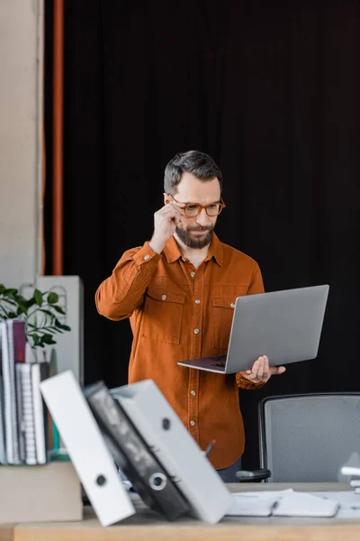 Homem de negócios barbudo sério ajustando óculos elegantes e olhando para laptop perto de pastas, cadernos e documentos na mesa de escritório em primeiro plano borrado — Fotografia de Stock