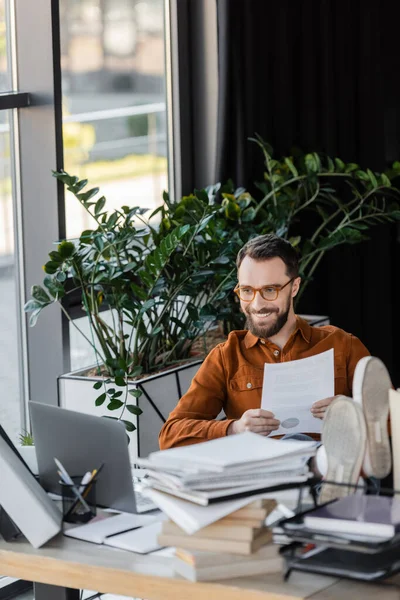 Hombre de negocios carismático y feliz en gafas con documento mientras está sentado con las piernas en el escritorio cerca de la computadora portátil, pila de cuadernos y plantas decorativas en la oficina - foto de stock