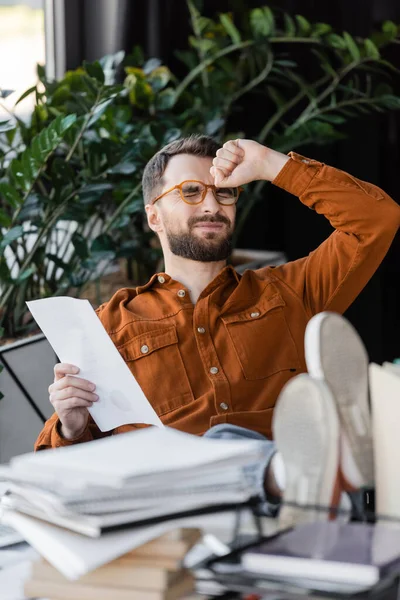 Ocupado e cansado empresário em óculos e camisa segurando documento enquanto sentado com as pernas na mesa e olhos fechados perto de pilha turva de cadernos na mesa de trabalho no escritório — Fotografia de Stock