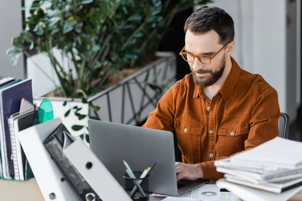 Homme d'affaires barbu positif à la mode lunettes et chemise travaillant sur ordinateur portable près pile de cahiers et dossiers avec des plantes décoratives sur fond flou dans le bureau — Stock Photo