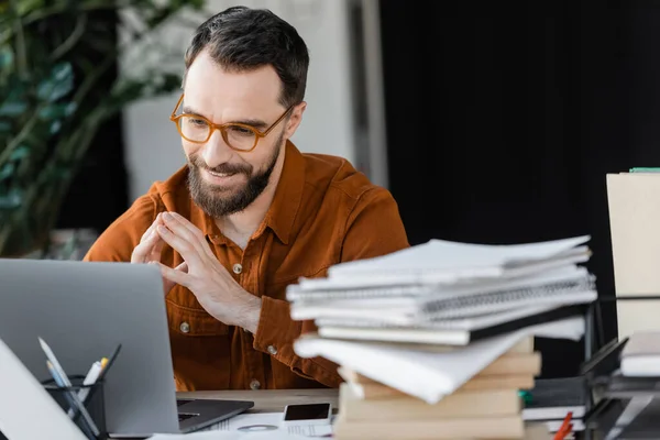Hombre de negocios barbudo con gafas y camisa con estilo mirando a la computadora portátil y sonriendo cerca de teléfono inteligente con pantalla en blanco y pila de cuadernos en primer plano borroso en la oficina - foto de stock