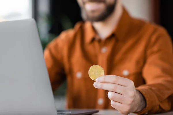 KYIV, UCRANIA - 18 de octubre de 2022: se centran en bitcoin de oro en la mano del exitoso hombre de negocios en camisa sentada cerca de la computadora portátil, fondo borroso, criptomoneda, finanzas - foto de stock