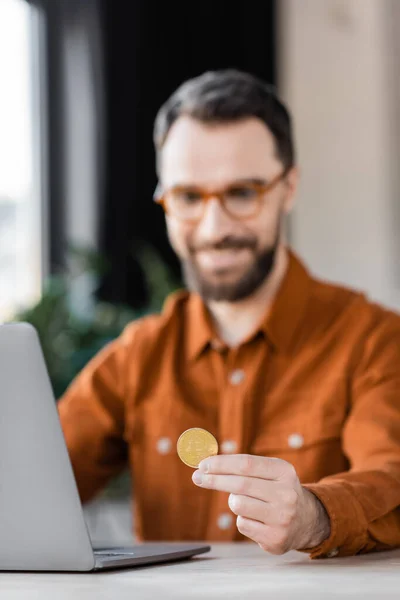 KYIV, UKRAINE - OCTOBER 18, 2022: smiling bearded businessman in eyeglasses and shirt sitting near laptop at workplace in office and holding golden bitcoin — Stock Photo