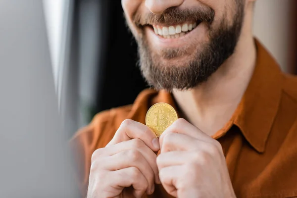 KYIV, UCRANIA - 18 de octubre de 2022: vista parcial del hombre de negocios barbudo y complacido sonriendo mientras sostiene el bitcoin dorado en un primer plano borroso en el cargo - foto de stock