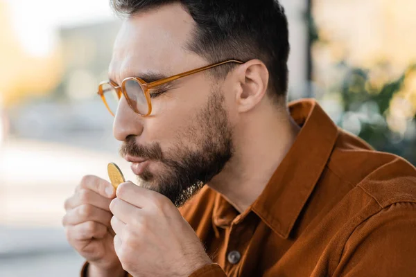 KYIV, UKRAINE - OCTOBER 18, 2022: bearded and brunette businessman in stylish eyeglasses and shirt, standing with closed eyes, kissing golden bitcoin in modern office on blurred background — Stock Photo