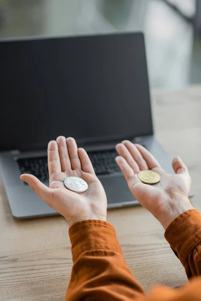 KYIV, UKRAINE - OCTOBER 18, 2022: cropped view of accomplished entrepreneur holding golden and silver bitcoins on open palms near laptop with blank screen on desk in office — Stock Photo