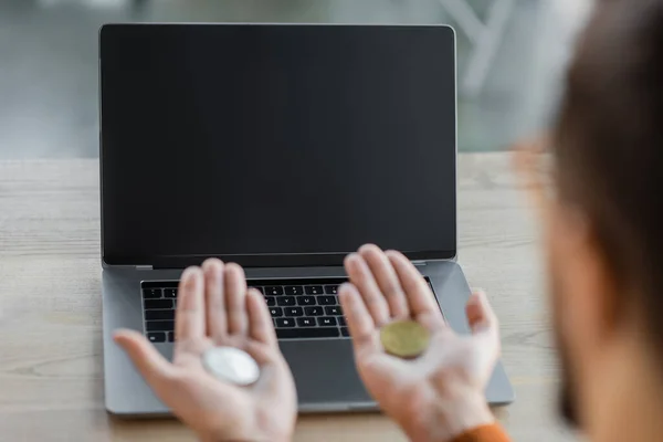 Abgeschnittene Ansicht eines verschwommenen Geschäftsmannes, der Bitcoins auf offenen Handflächen hält, während er in der Nähe eines Laptops mit leerem Bildschirm auf dem Schreibtisch im modernen Büro sitzt, verschwommener Vordergrund — Stockfoto
