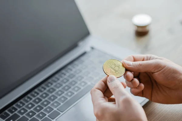 KYIV, UKRAINE - OCTOBER 18, 2022: cropped view of ambitious businessman holding golden bitcoin near laptop with blank screen on office desk, blurred background — Stock Photo