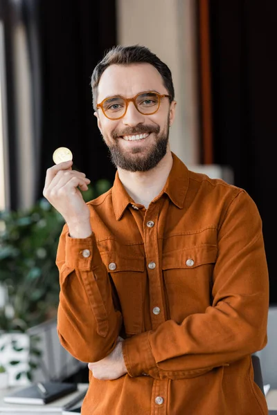KYIV, UKRAINE - OCTOBER 18, 2022: charismatic and bearded businessman in trendy eyeglasses and shirt holding golden bitcoin and smiling at camera in modern office on blurred background — Stock Photo