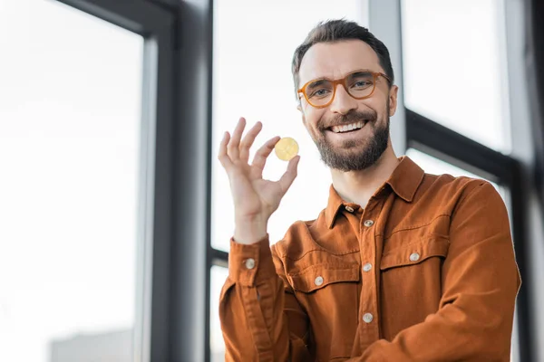KYIV, UKRAINE - OCTOBER 18, 2022: bearded businessman in fashionable shirt and eyeglasses holding golden bitcoin and smiling at camera near blurred windows in modern office on blurred background — Stock Photo