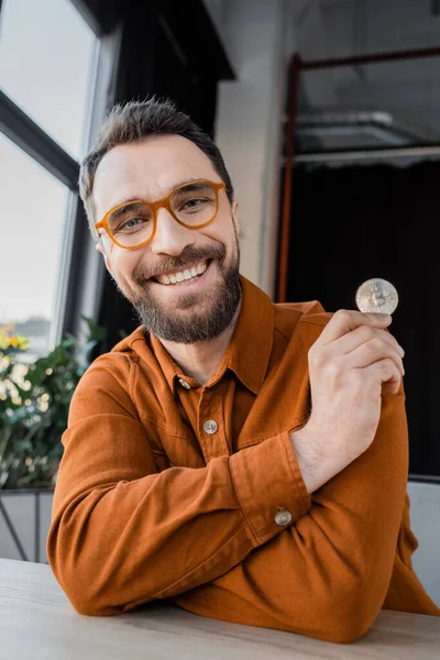 KYIV, UCRANIA - 18 de octubre de 2022: hombre de negocios lleno de alegría y barba en gafas de moda y camisa elegante celebración bitcoin plata y sonriendo a la cámara mientras está sentado en la oficina moderna - foto de stock