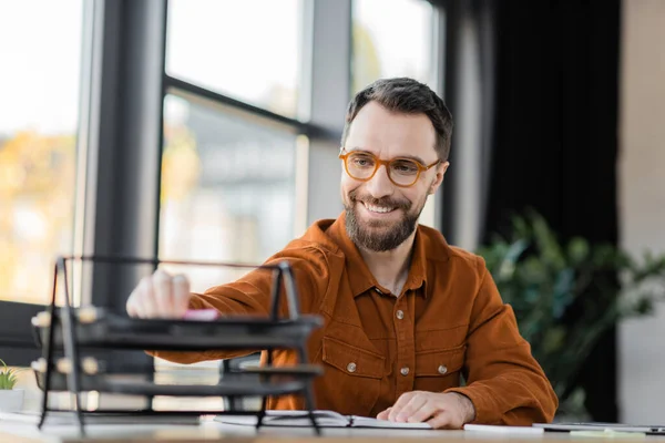 Successful and fashionable corporate manager in stylish shirt and eyeglasses sitting at workplace near notebook and blurred documents tray at workplace in office — Stock Photo