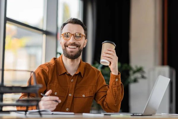 Charismatic bearded businessman in eyeglasses and shirt holding coffee to go and pen while smiling at camera next to notebook, laptop and smartphone in office, blurred foreground — Stock Photo