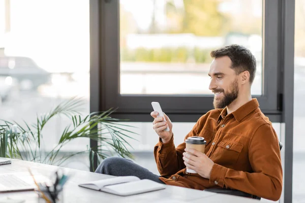Happy and successful bearded businessman with takeaway drink messaging on mobile phone near notebook, laptop, decorative plants and large windows in office — Stock Photo
