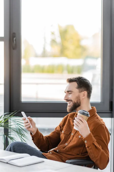Überglücklicher Geschäftsmann im trendigen Hemd, Kaffee in Pappbecher haltend und beim Sitzen am Arbeitsplatz neben großen Fenstern im modernen Büro auf das Handy blickend — Stockfoto
