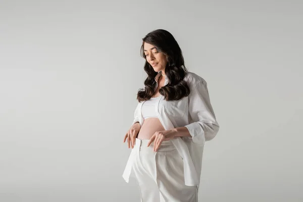 Очаровательная и счастливая беременная женщина с волнистыми волосами брюнетки, позирующими в белой рубашке, топе и штанах, улыбаясь изолированно на сером фоне, концепция моды материнства — стоковое фото