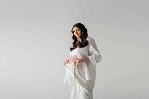 Happy future mother with wavy brunette hair posing in white fashionable clothes such as white shirt and pants isolated on grey background, trendy maternity concept — Stock Photo