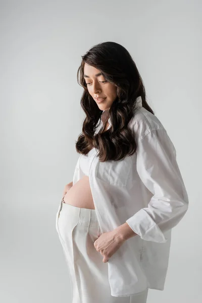 Очаровательная будущая мама в белой модной одежде, такой как белая рубашка, кукурузный топ и брюки, улыбающиеся изолированно на сером фоне, стильная концепция материнства, беременность — стоковое фото