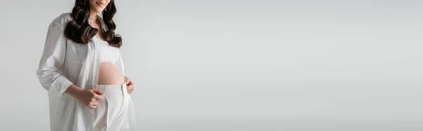 Vista cortada de mulher sorridente e grávida em camisa branca da moda e calças de pé isolado em fundo cinza, conceito de moda maternidade, banner — Fotografia de Stock